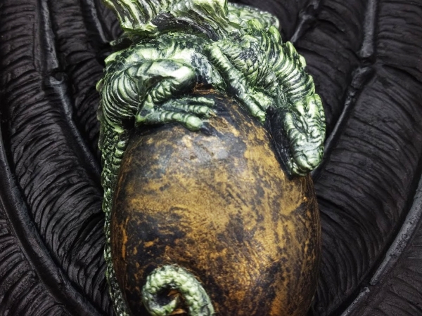 A green baby dragon rests on top of a golden egg, held gently by black wings.