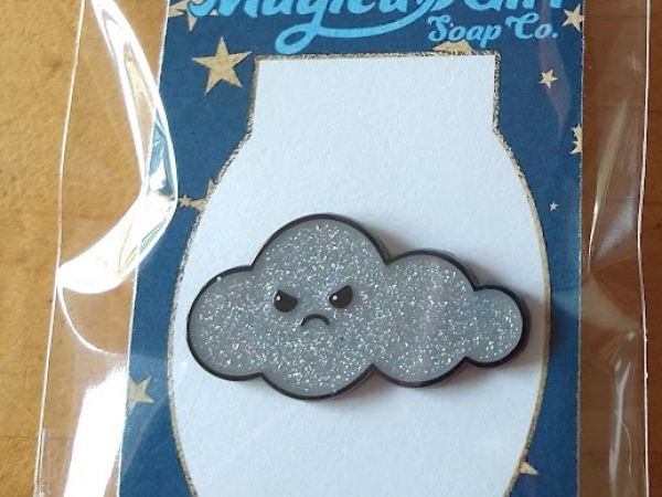 A photo of an enamel pin shaped like a cloud with a grumpy face.