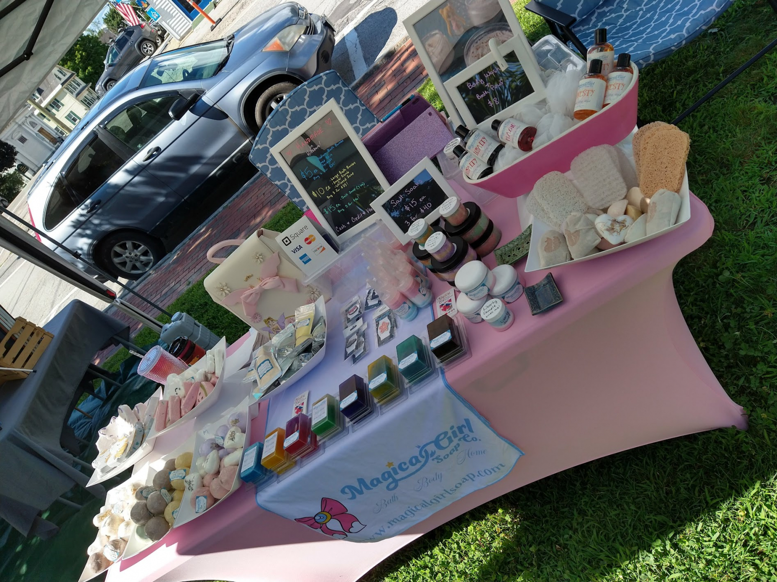 A photo of our setup at the summer market. There are a variety of soaps, scrubs, and bath bombs on a table covered by a pink tablecloth. 