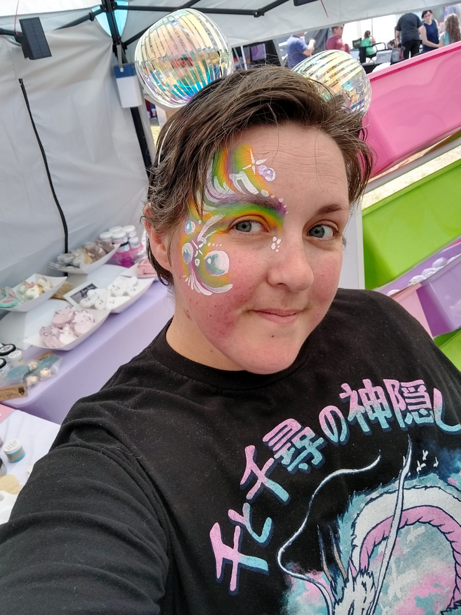 A person with short brown hair, bubbles on a headband, and rainbow face painting stands in the middle of our soap booth. <3